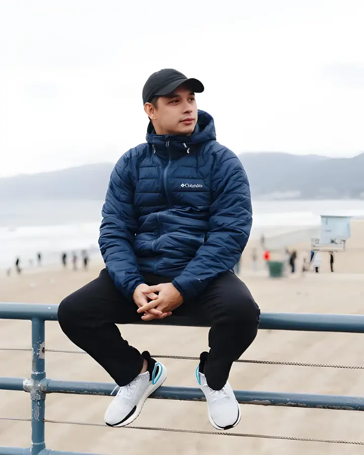 John Michael Lagumbay sitting on a railing by the beach, wearing a navy jacket and cap